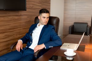 Confident businessman in blue suit seated at a desk with laptop, contemplating in office setting.